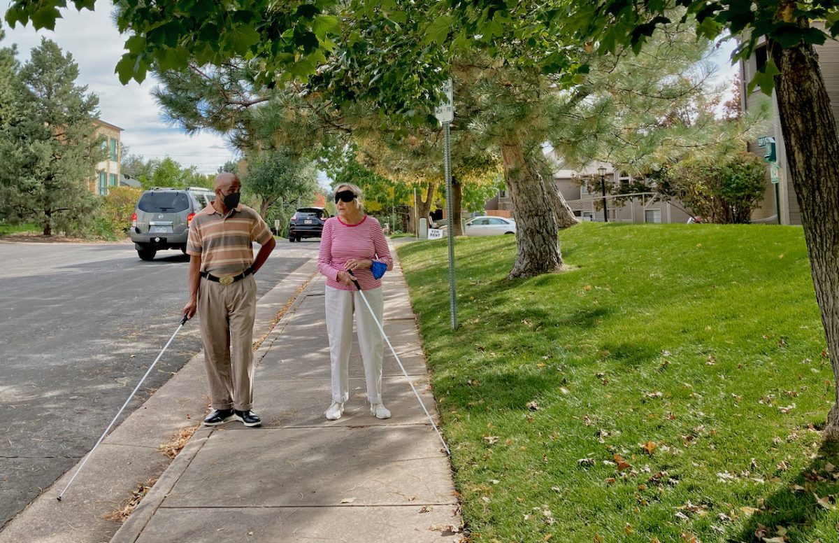 a blind instructor talks with his student, a senior woman as they walk with white canes along a residential street