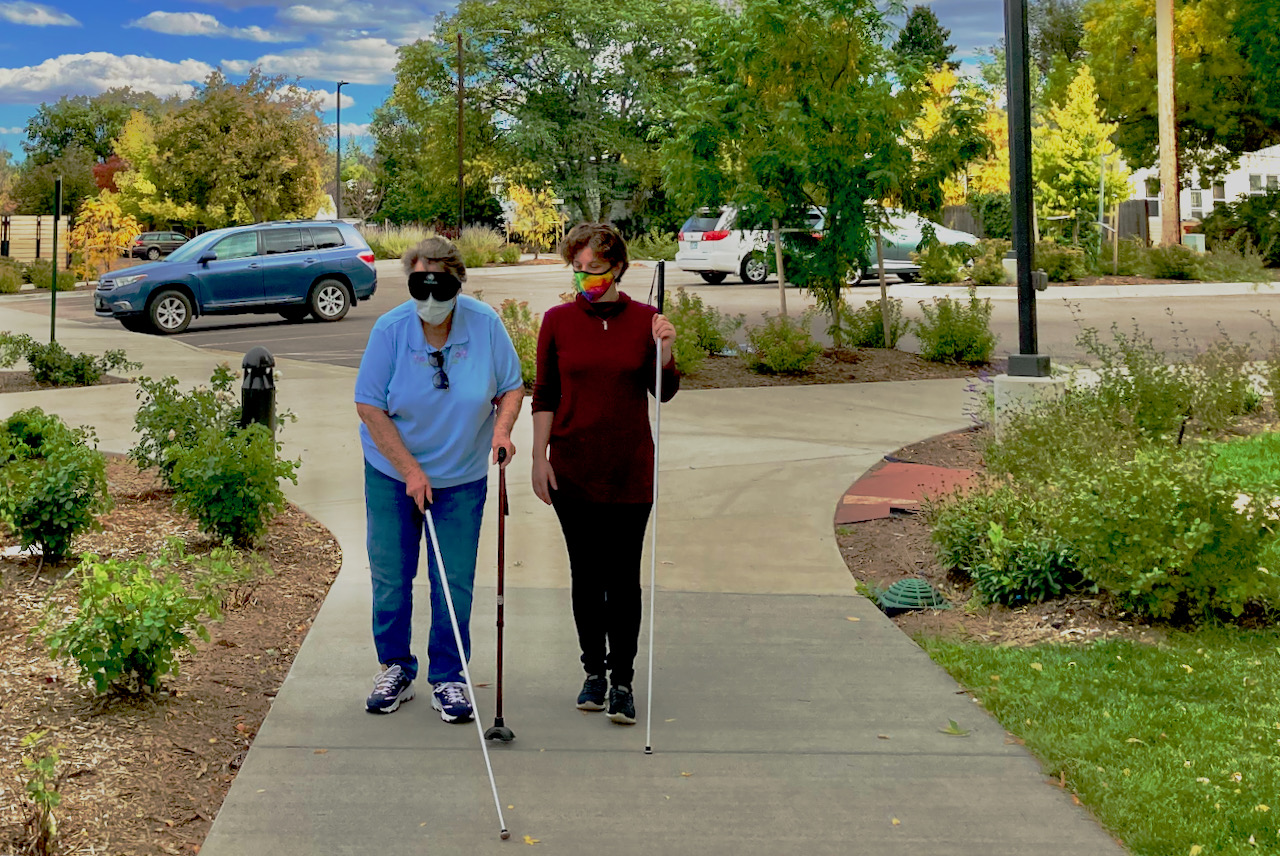 An older woman uses a support cane in her left hand and shorelines on the right with her white cane while her instructor observes