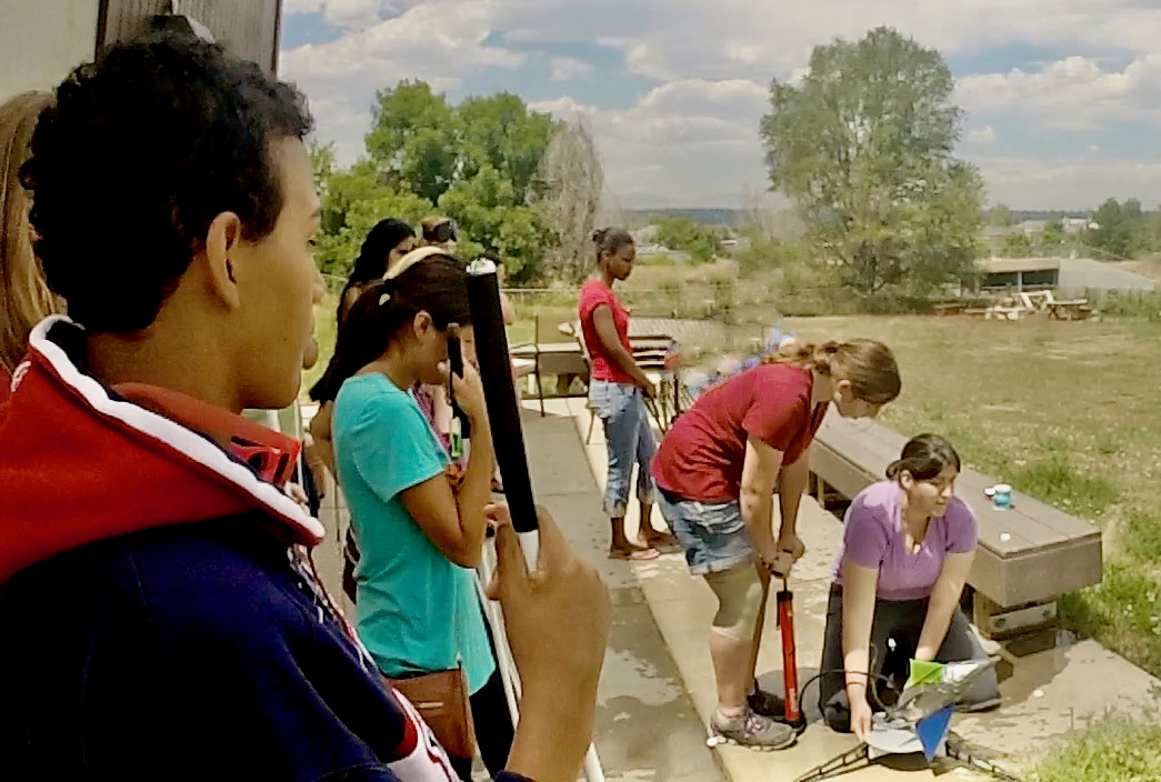 a high school student pumps air into a soda bottle rocket while the instructor kneels to steady the rocket for launch