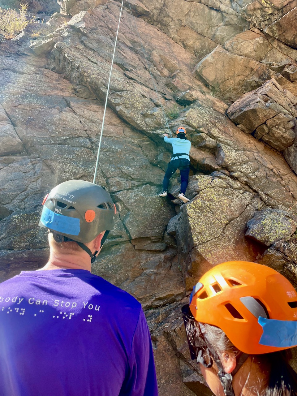 Ryan holds the belay rope for a student seen above on the rock. His t-shirt says “Nobody Can Stop You” in print and Braille.