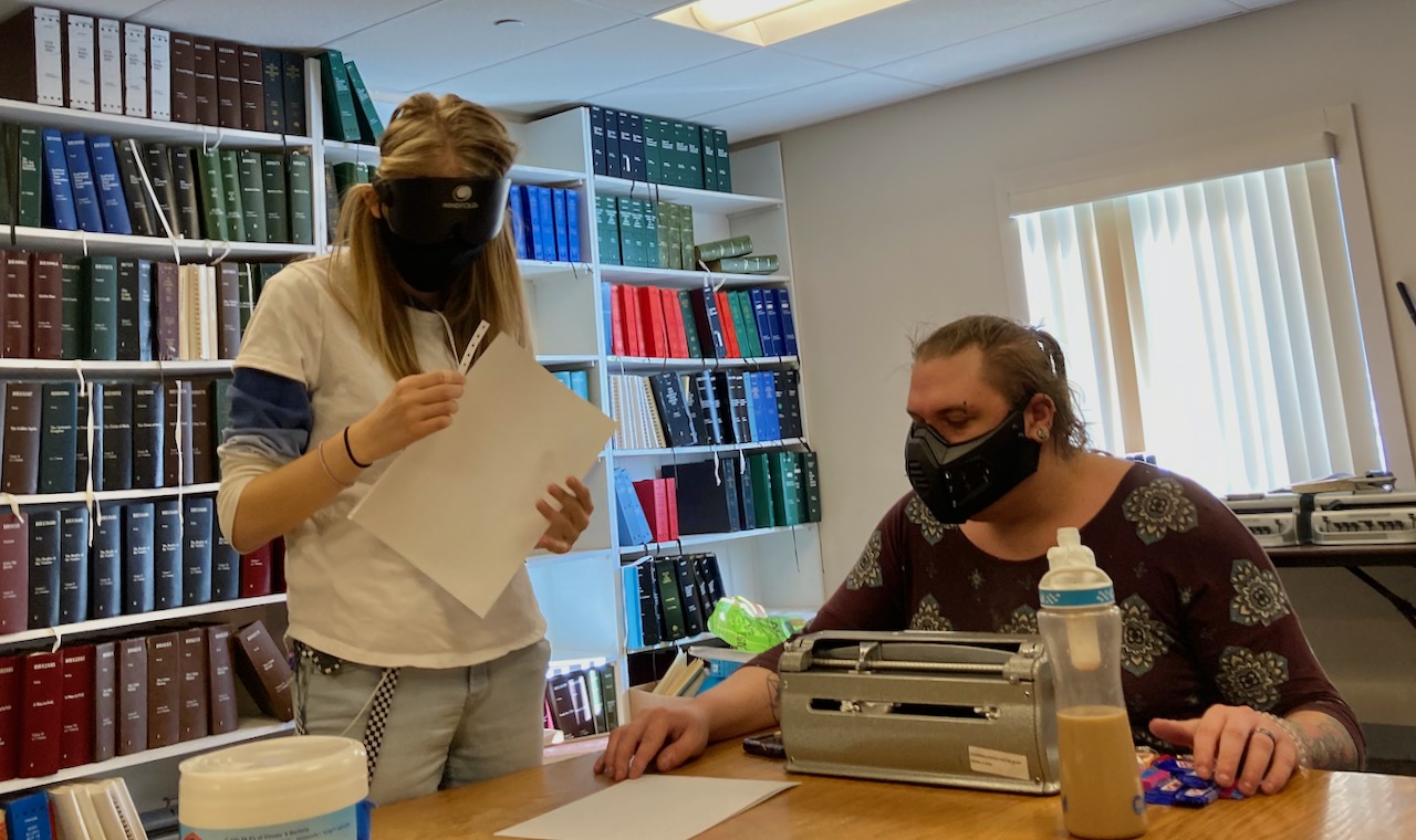 A young woman tears the tractor-feed edge from a piece of blank Braille paper, another student beside uses the Perkins Brailler.