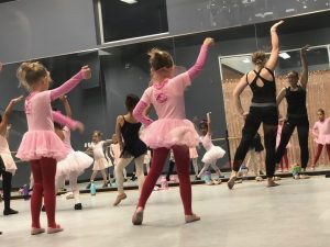 a class of five-year-olds in tights and tutus watches their instructor, her back to us but she is reflected in the dance studio mirror