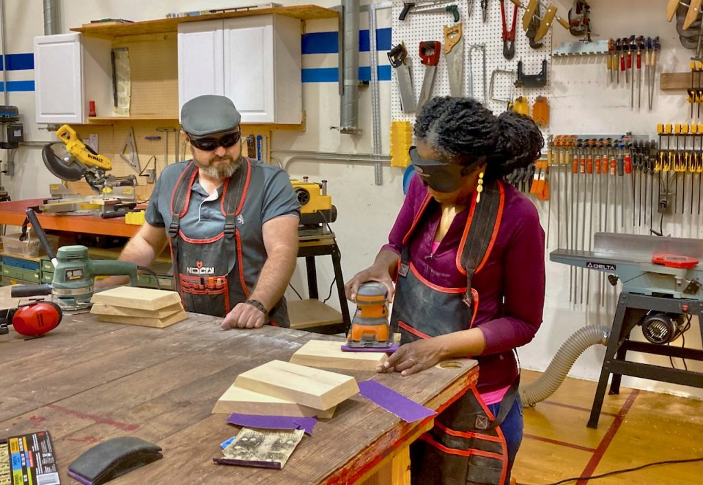 Rhonda uses an oscilating power sander in style, wearing a shop apron and long gold earrings!