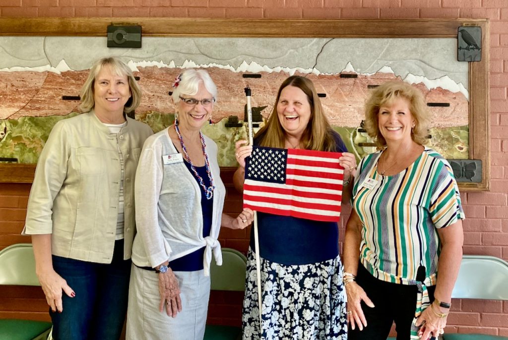 Four women stand together, smiling at the camera as the woman with a white cane displays the flag