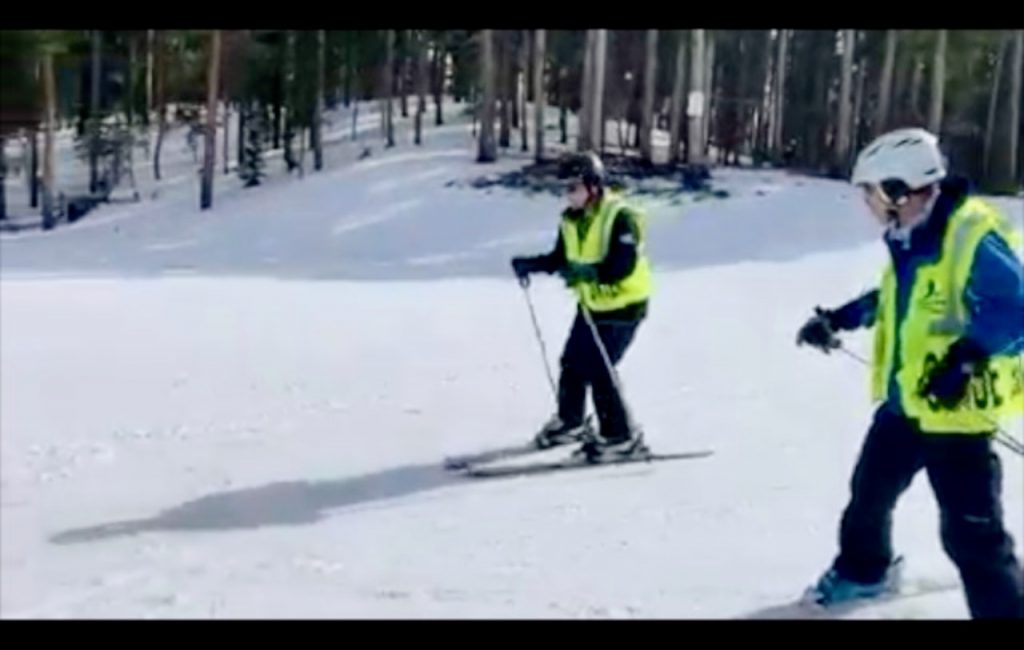 A man on skis wearing a "Blind Skier" vest moves down a snowy slope at the direction of a guide behind him