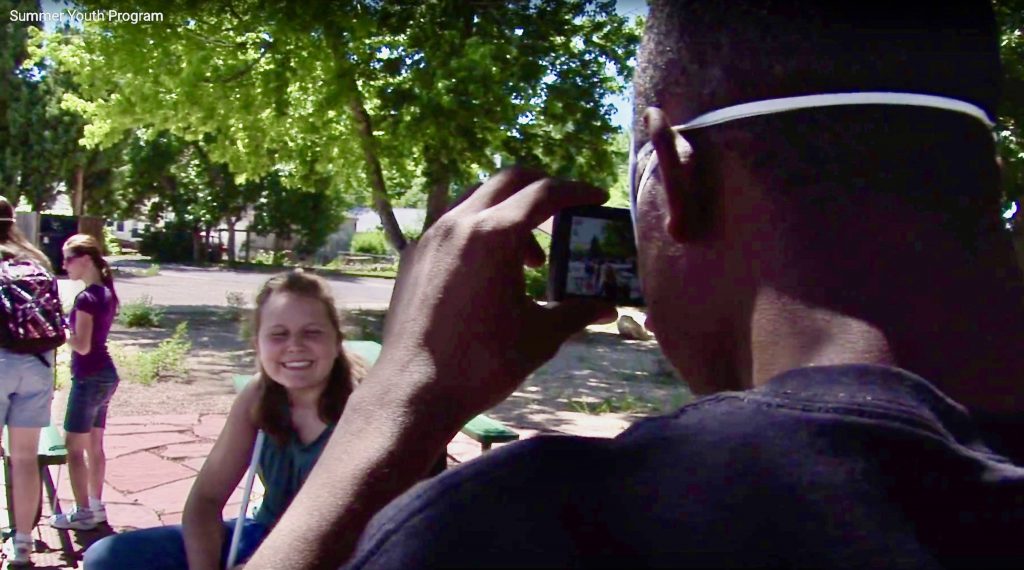 Group of Summer Youth at the Colorado Center picnic tables. One blind student takes a photo of another smiling student.