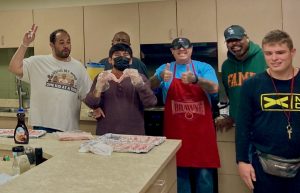 Six men face the camera across the kitchen island , some holding spatulas and one giving a double thumbs-up