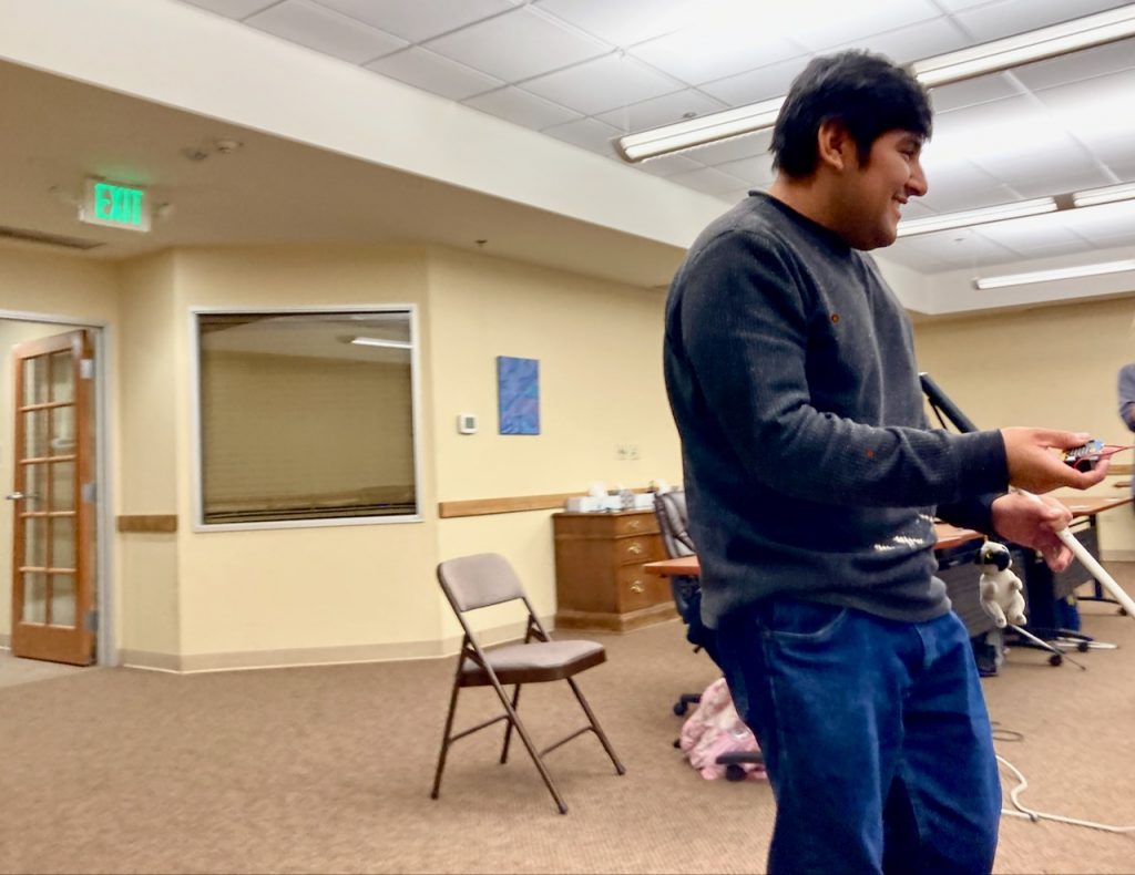 a young man with his cane holds out a small Microbit board as he explores the room for a quicker signal from the “lost” unit