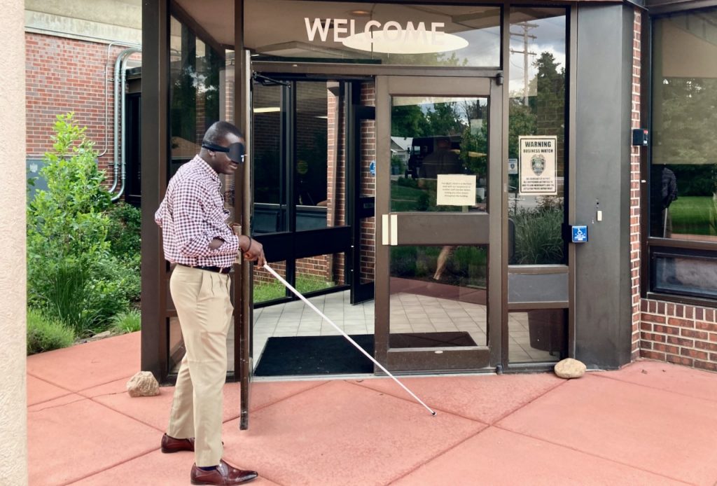 A man wearing learning shades finds the handle of the front door to the building, a “Welcome” sign above his head.