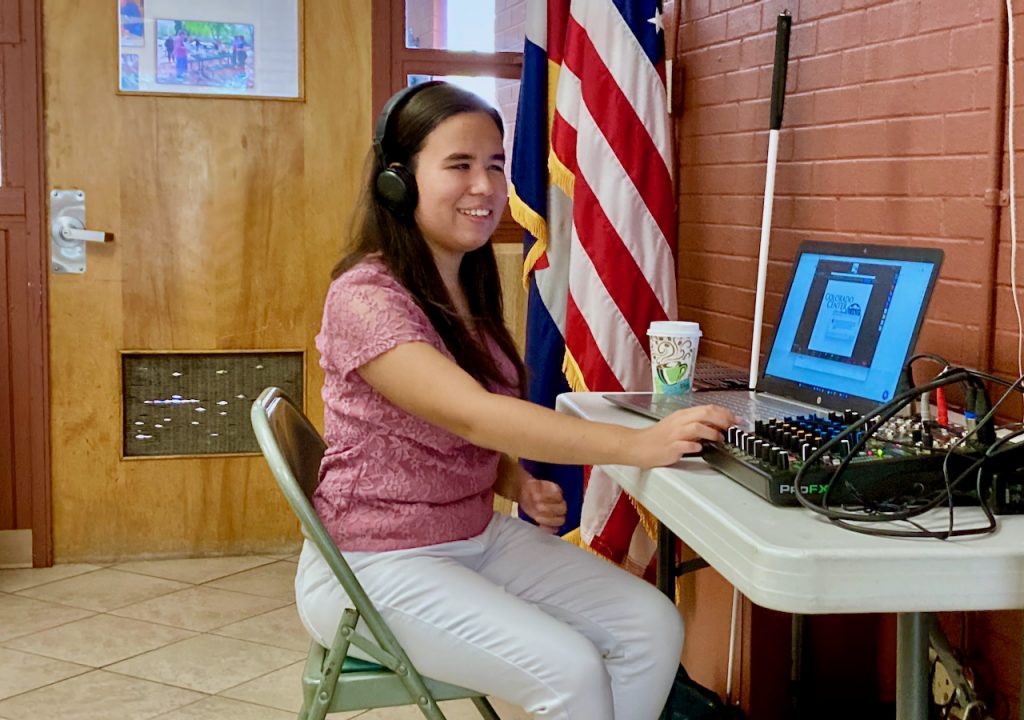 a smiling dark-haired young woman reaches for the mixing board, a laptop display shows the Colorado Center for the Blind logo on a Zoom screen