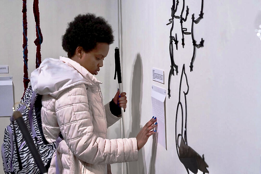 a young black woman holding a white cane reads the Braille title on the wall beside a tactile piece