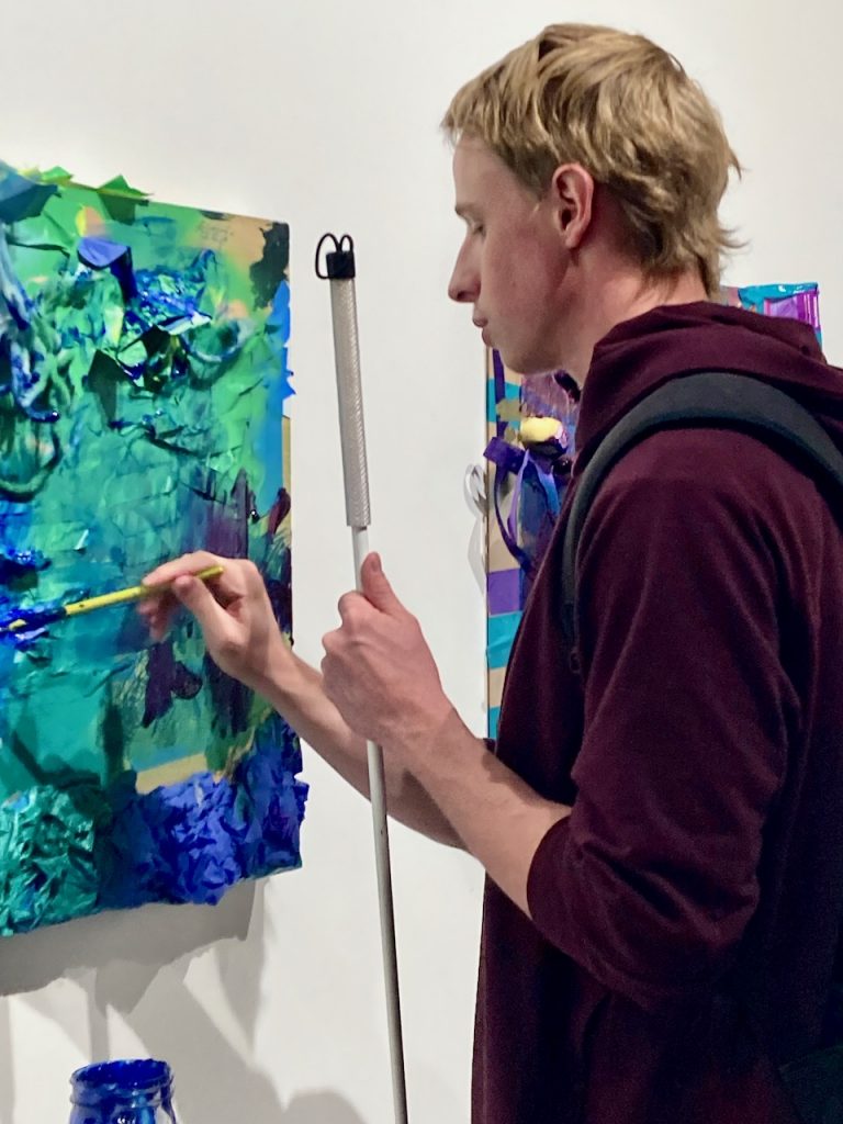 a young man holding a white cane swashes blue paint on an interactive art piece