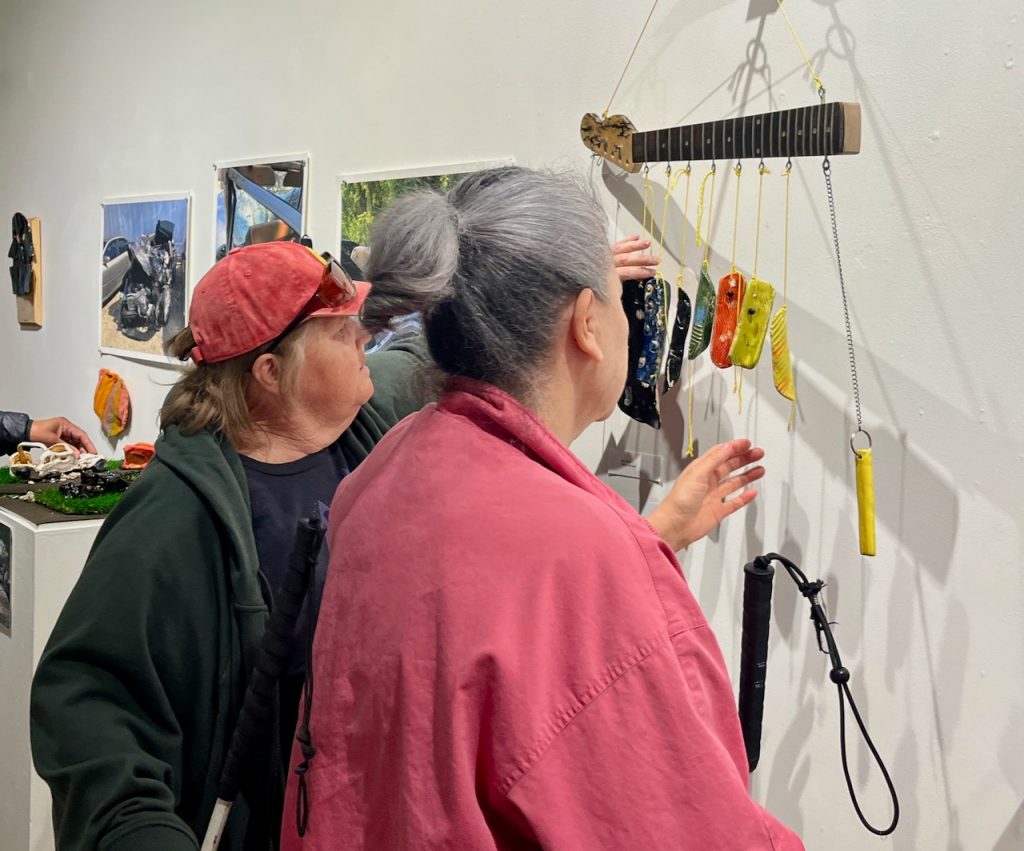 Two senior women holding white canes reach out to interact with colorful ceramic wind chimes dangling from a guitar neck