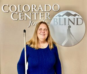 Julie stands and smiles while holding her white cane near the CCB logo in the Center Lobby