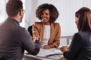 A nicely-dressed black woman reaches across an informational table to shake hands with a potential employer