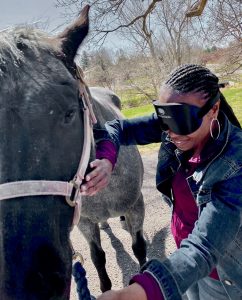 A woman with braided hair and learning shades holds a horse’s halter rope while she strokes the horse with her other hand