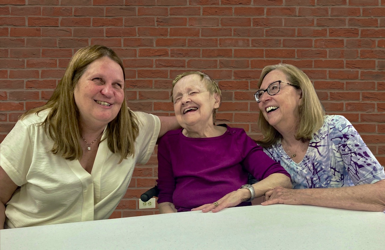 Two smiling professional women at a table lean in with their arms around a grinning, well-dressed older woman