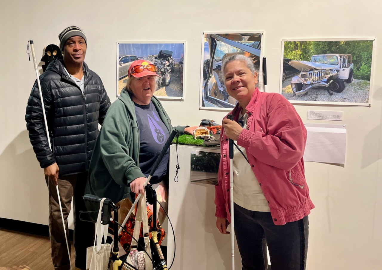 Several older blind people stand in front of a wall with art works