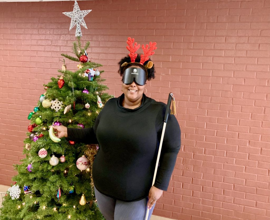 A smiling young woman wearing learning shades and reindeer antlers stands to the right of a lighted, colorfully decorated Christmas tree