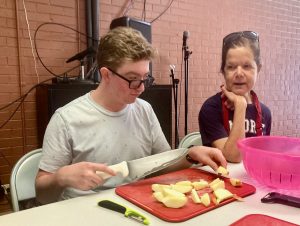 High school student Seth slices apples with a large knife while mentor Heather offers guidance.