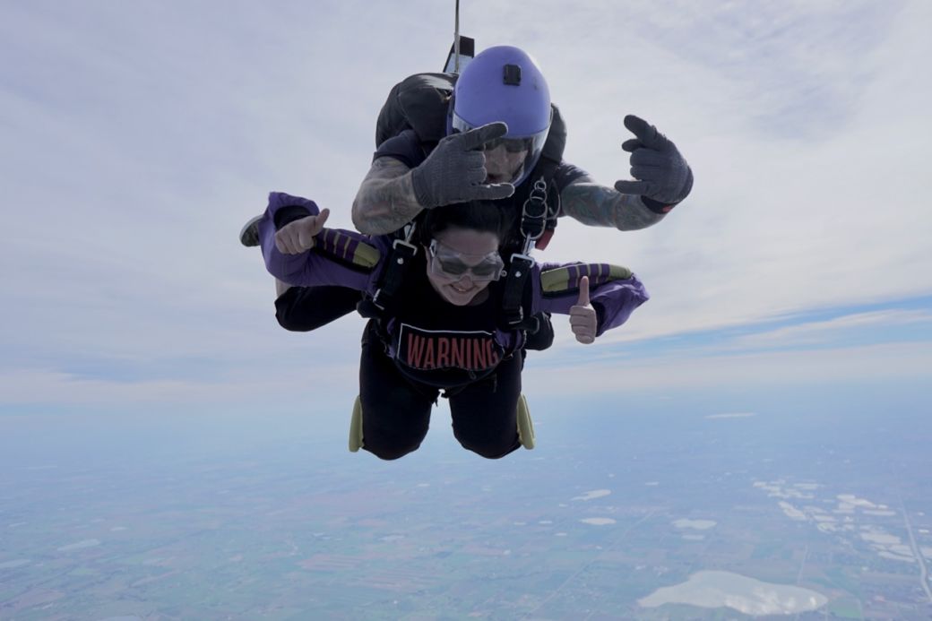 a grinning young woman strapped to her jump-master gives a double thumbs-up
