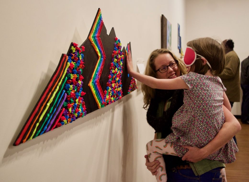 Katy holds up her daughter Zoey who is wearing sleepshades so she can touch ta tactile art piece of the mountains