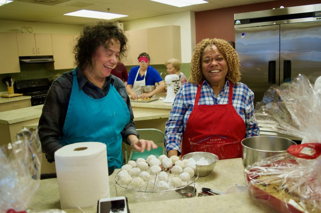 Blanca and Priscilla wearing festive colored aprons prepare a rack of home made powdered sugar cookies