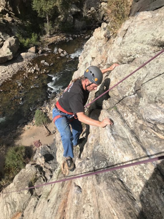 Looking down at Brad who is nearly 80 feet up the rock face. We see tall trees and the river far below.