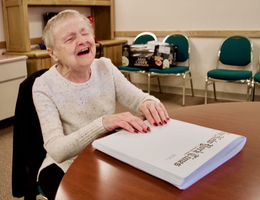 A small blind woman laughs as her hands rest on a Braille copy of the New York Times Weekly.