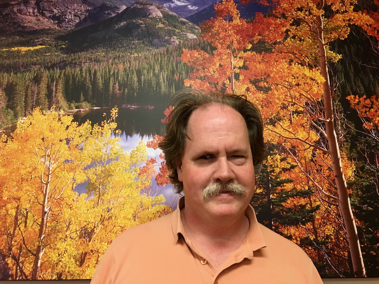 Chip Johnson standing in front of a colorful Colorado Mountain Lake landscape in the Small Conference Room